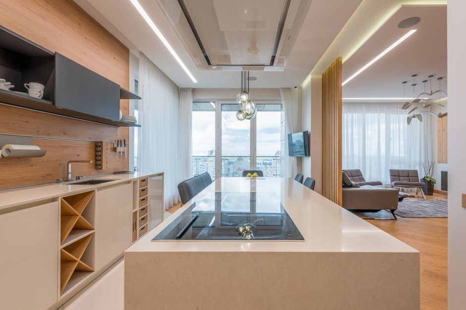 Shiny stone island counter with built stove top and white cabinets in light open plan kitchen with gray sofas in lounge area