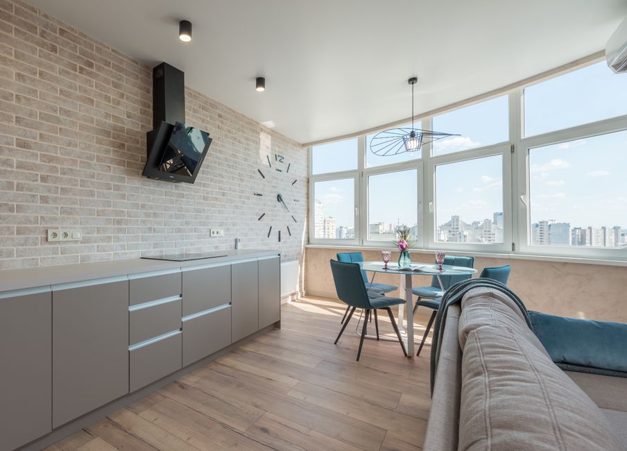 Interior design of new stylish studio consisting of spacious kitchen area near comfortable couch and dining zone with clock on brick wall and big windows